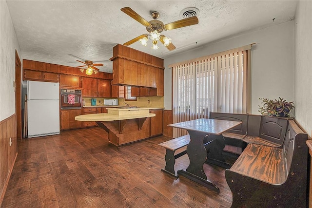 kitchen featuring black oven, wainscoting, freestanding refrigerator, brown cabinets, and dark wood-style floors