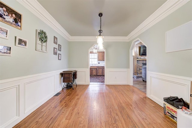 unfurnished dining area with arched walkways, a wainscoted wall, light wood-style flooring, ornamental molding, and a brick fireplace