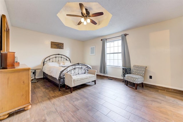 bedroom with a textured ceiling, ceiling fan, wood finished floors, baseboards, and a tray ceiling