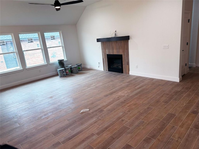 unfurnished living room with dark wood-style floors, a tiled fireplace, a ceiling fan, vaulted ceiling, and baseboards