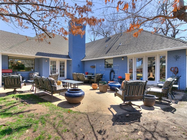 back of house with french doors, an outdoor living space with a fire pit, roof with shingles, brick siding, and a chimney