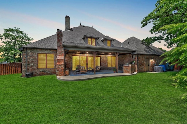 back of property at dusk with brick siding, fence, a lawn, a chimney, and a patio