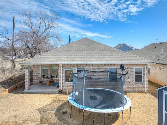 rear view of property with a trampoline, roof with shingles, a patio, brick siding, and fence