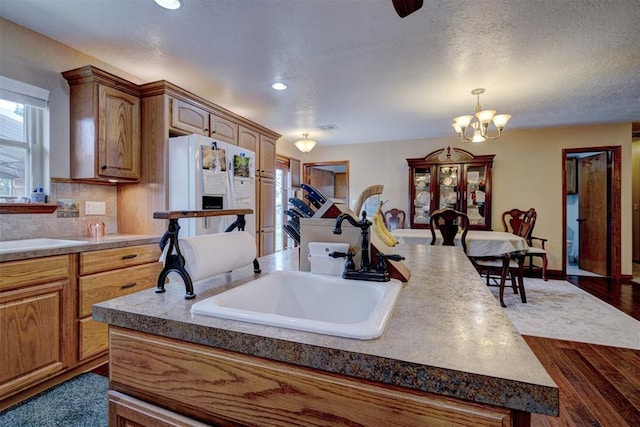 kitchen featuring white refrigerator with ice dispenser, dark wood-type flooring, a sink, a kitchen island with sink, and a notable chandelier