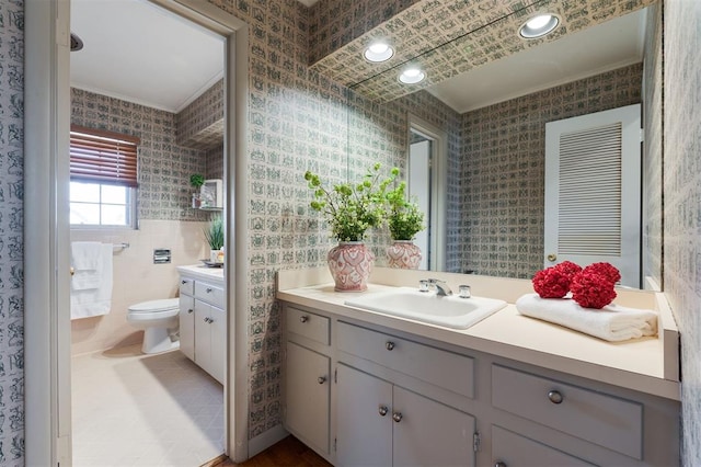 bathroom featuring wallpapered walls, toilet, crown molding, vanity, and tile walls