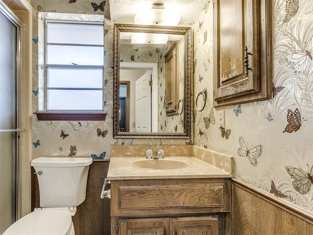 bathroom featuring toilet, a wainscoted wall, vanity, an enclosed shower, and wallpapered walls