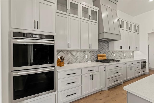 kitchen featuring custom exhaust hood, backsplash, double oven, white cabinetry, and gas cooktop