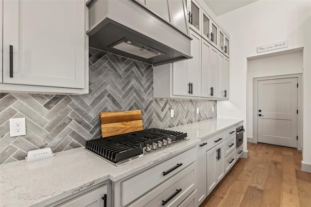 kitchen featuring stainless steel gas cooktop, premium range hood, white cabinetry, light wood-type flooring, and glass insert cabinets