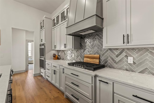 kitchen with premium range hood, appliances with stainless steel finishes, light wood-type flooring, tasteful backsplash, and glass insert cabinets