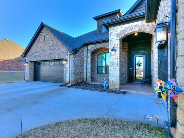 doorway to property featuring stone siding, roof with shingles, an attached garage, and concrete driveway