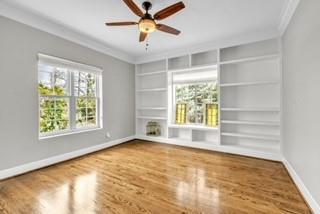 empty room with built in shelves, wood finished floors, a ceiling fan, baseboards, and ornamental molding