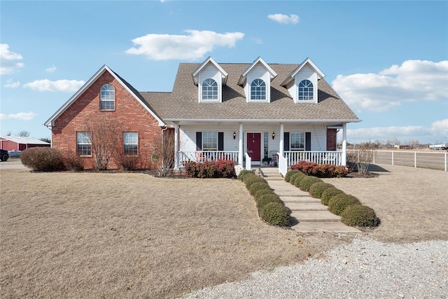 cape cod house featuring a porch, roof with shingles, brick siding, and fence