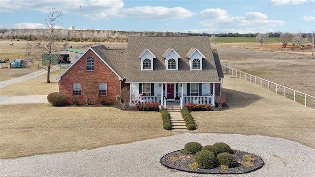 cape cod-style house with curved driveway, a rural view, fence, a porch, and brick siding