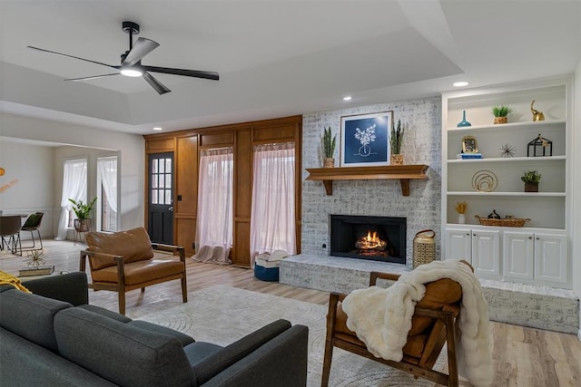 living area featuring light wood finished floors, ceiling fan, a brick fireplace, and a tray ceiling