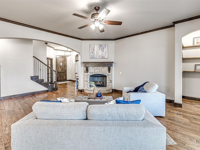 living area with arched walkways, ceiling fan, a stone fireplace, wood finished floors, and stairs