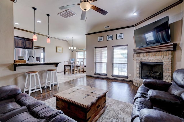 living area with a brick fireplace, ceiling fan with notable chandelier, visible vents, and ornamental molding