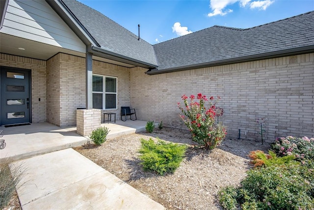 property entrance featuring covered porch, a shingled roof, and brick siding