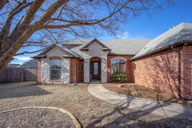 french provincial home featuring stone siding, a shingled roof, fence, and brick siding