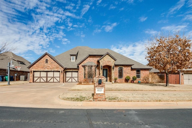 french country style house with an attached garage, brick siding, fence, driveway, and roof with shingles