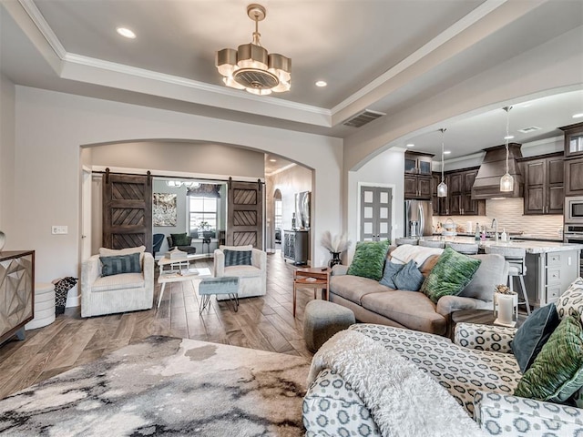 living room with arched walkways, a barn door, visible vents, a raised ceiling, and crown molding