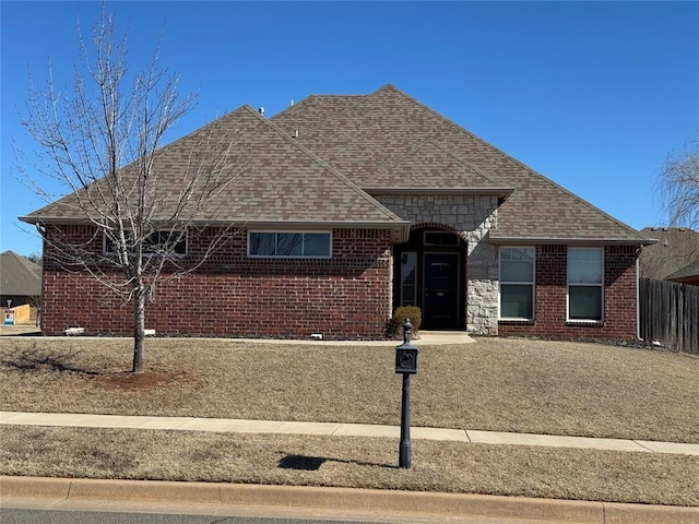 view of front facade featuring stone siding, a shingled roof, fence, and brick siding