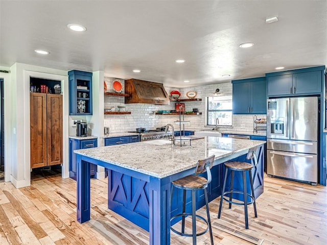 kitchen featuring blue cabinets, appliances with stainless steel finishes, light wood-type flooring, custom exhaust hood, and open shelves