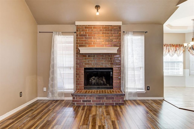 unfurnished living room featuring a chandelier, a brick fireplace, wood-type flooring, and baseboards