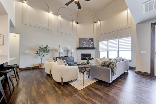 living area with visible vents, a ceiling fan, a towering ceiling, dark wood-style floors, and a stone fireplace