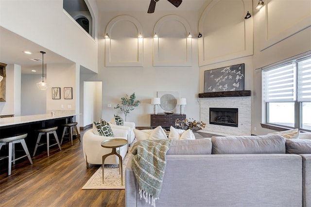 living room featuring dark wood-style flooring, ceiling fan, a stone fireplace, and a high ceiling