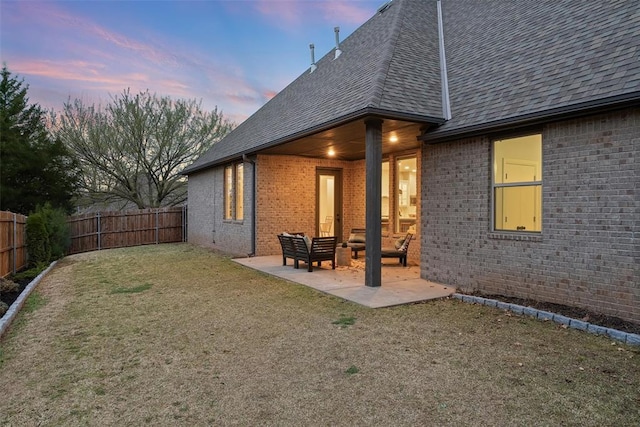 back of house at dusk with a shingled roof, a fenced backyard, brick siding, and a patio area