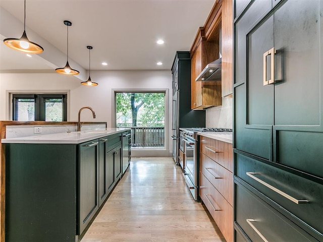 kitchen with light wood finished floors, a sink, wall chimney range hood, stainless steel appliances, and a kitchen island with sink