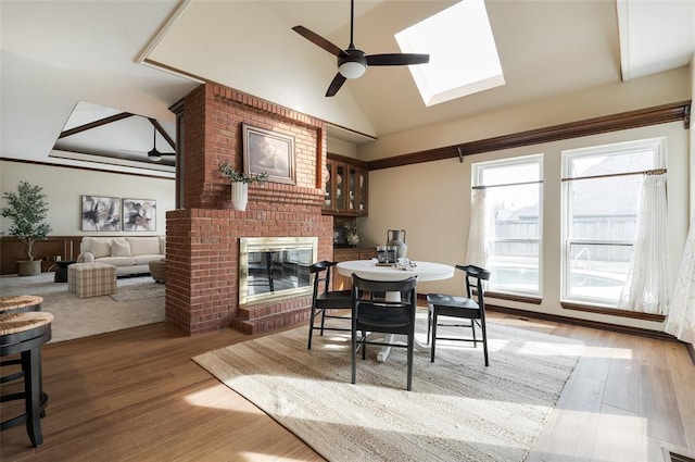 dining area featuring visible vents, light wood-style flooring, lofted ceiling with skylight, a ceiling fan, and a brick fireplace