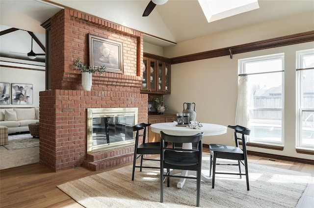 dining room featuring a brick fireplace, vaulted ceiling with skylight, light wood-style floors, and a wealth of natural light