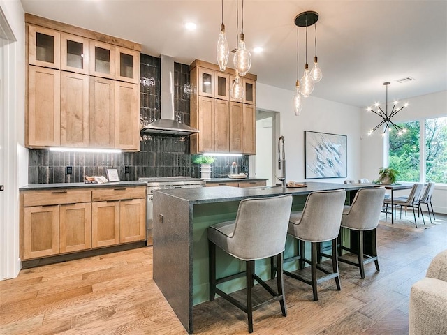 kitchen with high end stainless steel range, decorative backsplash, an island with sink, light wood-type flooring, and wall chimney exhaust hood