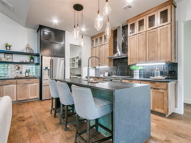 kitchen with light wood-style flooring, dark countertops, wall chimney range hood, and stainless steel fridge with ice dispenser