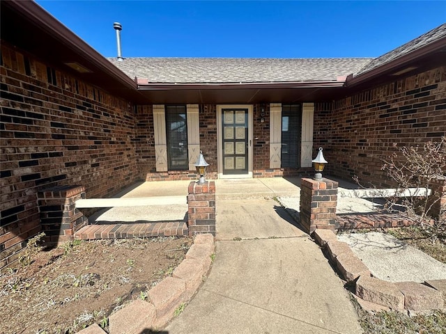 view of exterior entry with covered porch, roof with shingles, and brick siding