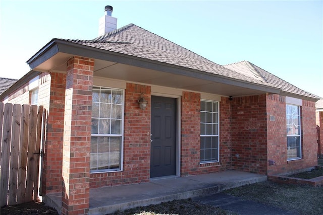 doorway to property featuring covered porch, brick siding, a shingled roof, fence, and a chimney