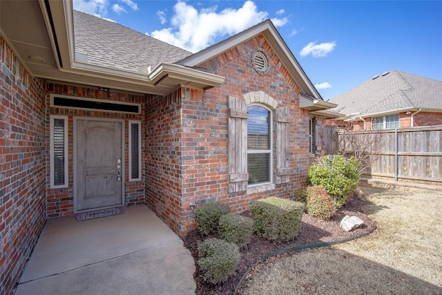 entrance to property featuring a shingled roof, fence, and brick siding