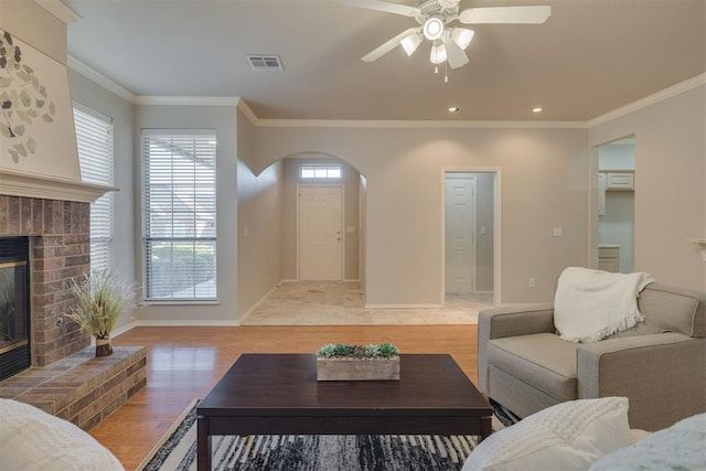 living area featuring visible vents, light wood-style flooring, arched walkways, ornamental molding, and a brick fireplace