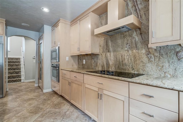 kitchen with wall chimney range hood, black electric stovetop, light brown cabinetry, and oven