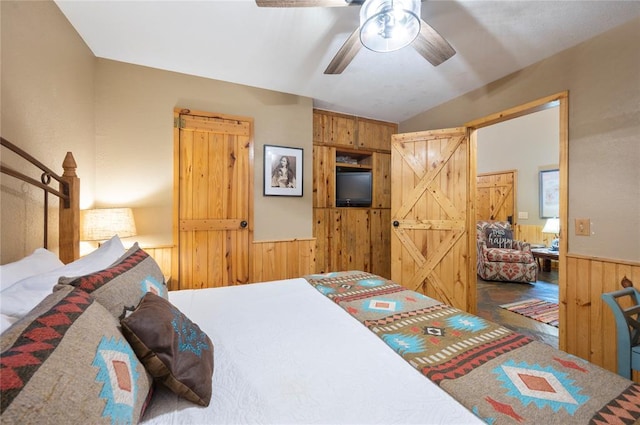 bedroom featuring a barn door, a ceiling fan, wainscoting, vaulted ceiling, and wood walls