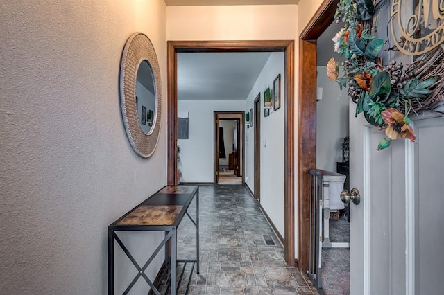 hall featuring stone finish floor, visible vents, a textured wall, and baseboards