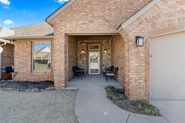 view of exterior entry with a garage, brick siding, and roof with shingles