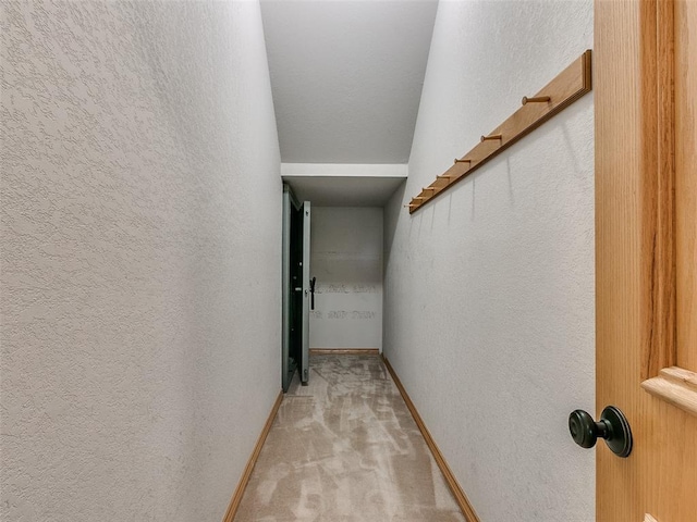 hall featuring baseboards, a textured wall, and light colored carpet