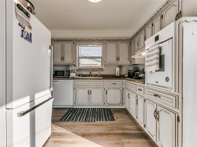 kitchen with light wood finished floors, dark countertops, a sink, white appliances, and under cabinet range hood