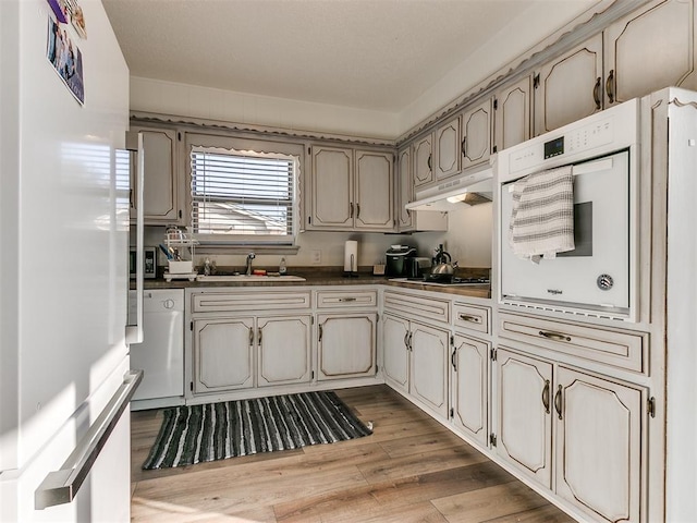 kitchen featuring under cabinet range hood, white appliances, a sink, light wood finished floors, and dark countertops