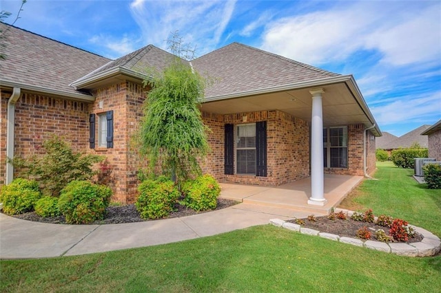 entrance to property featuring brick siding, central AC unit, a shingled roof, and a yard