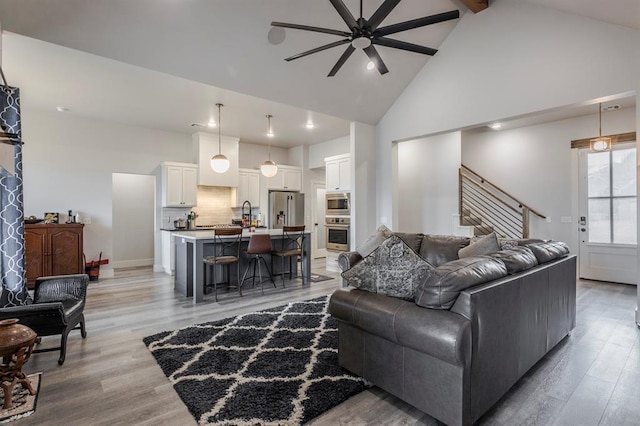 living area with stairway, beam ceiling, light wood-style floors, high vaulted ceiling, and a ceiling fan