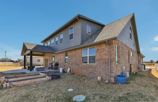 back of house featuring a patio, a fire pit, brick siding, roof with shingles, and a lawn