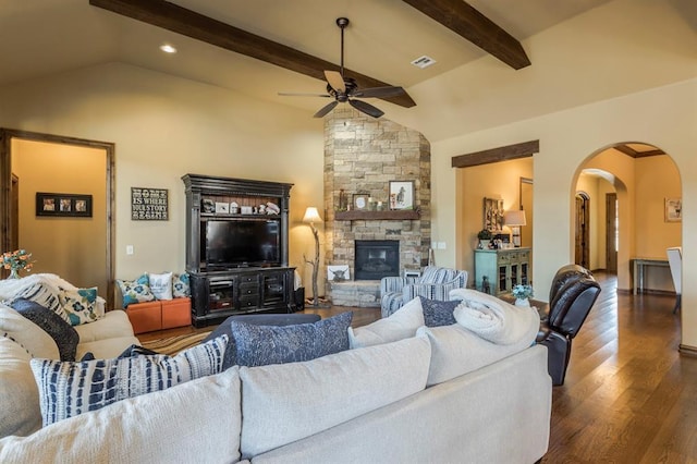 living area with arched walkways, vaulted ceiling with beams, ceiling fan, a stone fireplace, and wood finished floors